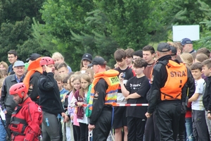 Na zdjęciu strażak w kombinezonie do pływania tłumaczy zebranej młodzieży jak się bezpiecznie zachowywać nad wodą.