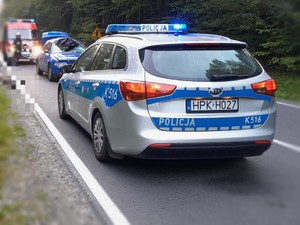 Na zdjęciu,  na pierwszym planie poznakowany radiowóz policyjny, w tle samochód osobowy koloru niebieskiego z widocznymi uszkodzeniami, wóz strażacki oraz las.