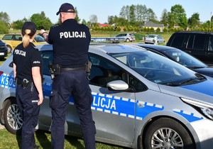 Na zdjęciu umundurowany policjant i policjantka w tle oznakowany samochód policji.