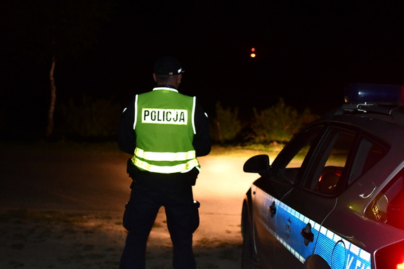 Na zdjęciu noc policjant w umundurowaniu służbowym z kamizelką odblaskową i latarką i radiowóz.