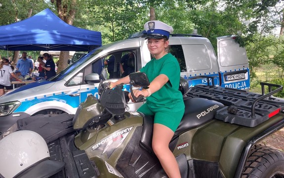 Na zdjęciu dziewczynka na quadzie w policyjnej czapce w tle oznakowany radiowóz.
