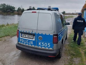 Na tle rzeki znajduje się oznakowany radiowóz oraz umundurowany policjant