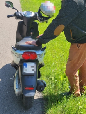 Na zdjęciu, po lewej stronie motorower, przy którym znajduje się mężczyzna, trzymający ręce na pojeździe.