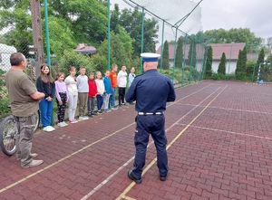 Na zdjęciu policjant w umundurowaniu przeprowadza egzamin na kartę rowerową, w tle uczniowie wraz z opiekunem.