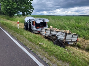 Na zdjęciu w rowie samochód osobowy koloru szarego z przyczepką wypełnioną drzewem. Widoczne uszkodzenia koła lewego przyczepki.