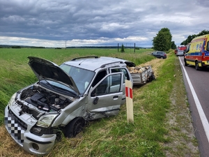 Na zdjęciu w rowie pojazd koloru szarego z widocznymi uszkodzeniami maski przedniej oraz po stronie lewej pojazdu, w tle przyczepka wypełniona drzewem, karetka pogotowia ratunkowego, wóz straży pożarnej oraz inne samochody.