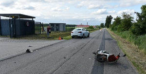 Na zdjęciu miejsce zdarzenia drogowego.  Po prawej stronie zdjęcia motorower koloru czerwono szarego przewrócony na lewy bok, w tle samochód osobowy koloru białego. Oba pojazdy znajdują się na drodze, na której leżą elementy pojazdów.