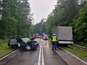 Na zdjęciu funkcjonariusz ruchu drogowego w umundurowaniu służbowym i w kamizelce odblaskowej wykonuje czynności w miejscu zderzenia dwóch pojazdów. Po stronie lewej samochód osobowy koloru czarnego z widocznymi uszkodzeniami części czołowej pojazdu oraz elementów strony prawej samochodu. Po stronie prawej naczepa samochodu ciężarowego, w tle strażacy,  wozy strażackie, samochody które zatrzymały się przed miejscem zdarzenia.