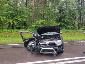 Na zdjęciu samochód osobowy koloru czarnego z uszkodzeniami powstałymi w trakcie zderzenia pojazdów. Samochód posiada wyrwane koło lewe, uszkodzenia strony lewej pojazdu, uszkodzenia części czołowej, maska pojazdu uniesiona do góry, wygięta.