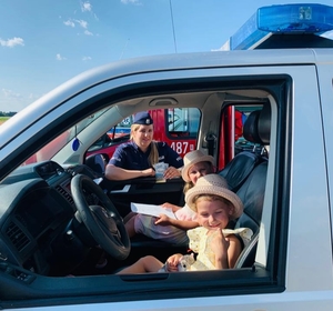 Na zdjęciu dzieci siedzące w oznakowanym radiowozie, w tle policjantka w umundurowaniu służbowym.