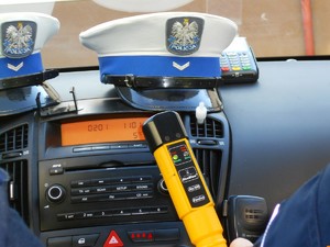 Na zdjęciu wewnątrz radiowozu, na podszybiu leżą dwie czapki służbowe policjantów ogniwa ruchu drogowego, obok terminal płatniczy, poniżej policjant trzyma w ręce urządzenie do sprawdzania stanu trzeźwości.