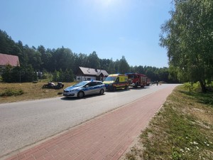 Zdjęcie przedstawia miejsce zdarzenia drogowego w miejscowości Ruda Rużaniecka. Na zdjęciu znajdują się radiowóz Policji, Karetka Pogotowia oraz Wóz Strażacki. W tle leży motocykl, oraz pracujący funkcjonariusze Komendy Policji w Lubaczowie oraz Strażacy.