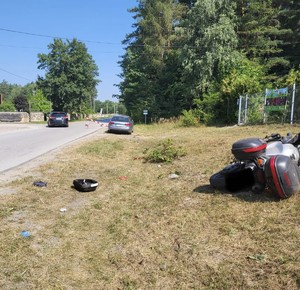 Zdjęcie przedstawia miejsce zdarzenia drogowego , widnieje motocykl koloru szarego, samochód osobowy koloru srebrnego oraz czarne auto osobowe. W tle las.