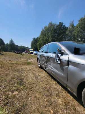 Zdjęcie przedstawia miejsce zdarzenia drogowego. Po prawej stronie pojazd koloru szarego który brał udział w zdarzeniu drogowym, w tle stoi radiowóz Policja, karetka pogotowia oraz Wóz strażacki.