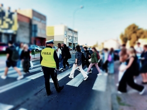 Na zdjęciu na pierwszym planie umundurowany policjant ruchu drogowego czuwa nad bezpieczeństwem osób przechodzących przez przejście dla pieszych.