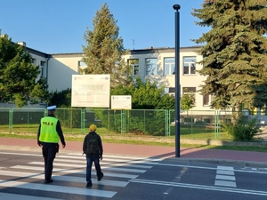 Na zdjęciu policjant w umundurowaniu służbowym pomaga przejść przez przejście dla pieszych uczniowi szkoły podstawowej.