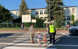 Na zdjęciu policjant w umundurowaniu służbowym przeprowadza przez przejście dla pieszych dzieci wraz z ojcem.