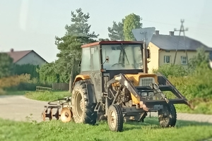 Na pierwszym planie ciągnik rolniczy z podpiętym pługiem. W tle zabudowania gospodarcze.