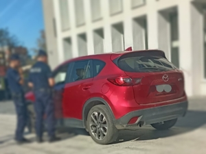 Na zdjęciu policjanci w umundurowaniu służbowym w trakcie kontroli drogowej samochodu osobowego koloru czerwonego.