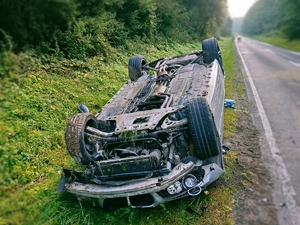 Na pierwszym planie zdjęcia pojazd marki BMW, który leży w rowie na dachu. Za zdjęciu uszkodzenia na całej powierzchni pojazdu. W tle  droga i otaczający ją las..