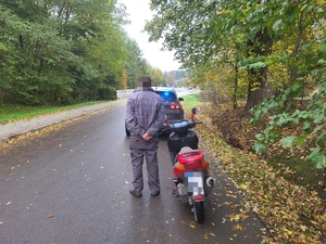 Na pierwszym planie zdjęcia mężczyzna w stroju roboczym stoi obok motoroweru w kolorze czerwono-czarnym. Na drugim planie nieoznakowany radiowóz policyjny. W tle droga asfaltowa oraz drzewa po lewej i prawej stronie drogi.