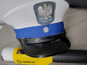 Na zdjęciu czapka służbowa policjanta ruchu drogowego z otokiem w kolorze niebieskim na którym widnieje stopień młodszego aspiranta policji. Czapka ma daszek w kolorze czarnym. Czapka jest ułożona na urządzeniu do sprawdzania stanu trzeźwości Promiler, który ma nakładkę w kolorze białym, trzon urządzenia jest w kolorze żółtym natomiast uchwyt w kolorze czarnym. Czapka i urządzenie do pomiaru stanu trzeźwości są ułożone na biurku.