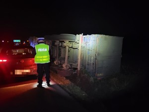 Na zdjęciu  na pierwszym planie policjant w umundurowaniu służbowym, stoi tyłem, obok niego po prawej stronie naczepa ciągnika leżąca w rowie, po stronie lewej nieoznakowany radiowóz policyjny z włączonymi sygnałami świetlnymi.