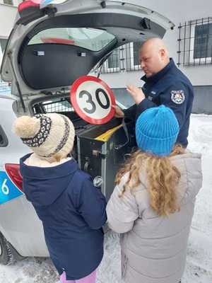 Na zdjęciu policjant w umundurowaniu służbowym stoi przy oznakowanym radiowozie i pokazuje dzieciom wyposażenie radiowozu.