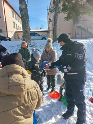 Zdjęcie przedstawia jak funkcjonariusz Policji rozdaje uczniom ulotki reklamowe dotyczące konkursu Śnieżny Delakog.