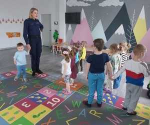 Fotografia przedstawia policjantkę w umundurowaniu służbowym, która wraz z przedszkolakami ćwiczy bezpieczne przechodzenie przez jezdnię.