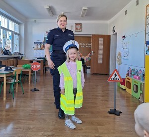 Na zdjęciu policjantka w umundurowaniu służbowym oraz dziewczynka ubrana w czapkę policyjną i kamizelkę odblaskową.