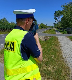 Na zdjęciu policjant w umundurowaniu służbowym i w kamizelce odblaskowej dokonujący pomiaru prędkości.