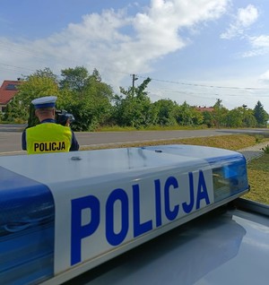 Na pierwszym planie galeryjka znajdująca się na radiowozie, w tle policjant w umundurowaniu służbowym dokonujący kontroli prędkości oraz droga.