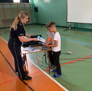 Na zdjęciu policjantka w umundurowaniu służbowym przekazująca ulotki dwóm chłopakom, w tle ściany hali sportowej.