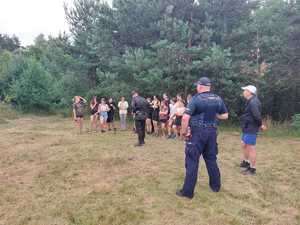 W tle znajduje się las a na pierwszym planie policjant w umundurowaniu służbowym, strażacy oraz uczestnicy spotkania.