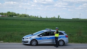 Na zdjęciu policjant w umundurowaniu służbowym stojący przy radiowozie w którym siedzi policjantka w umundurowaniu służbowym w tle pola.