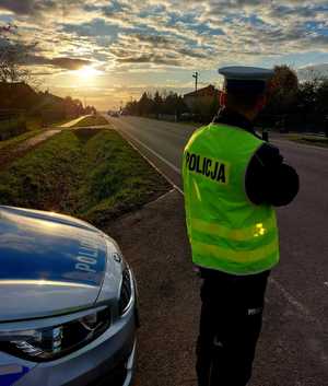 Na zdjęciu umundurowany policjant stojący obok radiowozu dokonujący pomiaru prędkości.