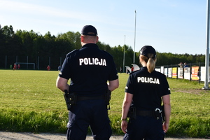 Na tle boiska sportowego, gdzie odgrywa się mecz piłki nożnej stoi policjant z policjantką w umundurowaniu służbowym.