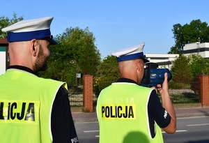 Na zdjęciu dwóch policjantów w umundurowaniu służbowym w kamizelkach odblaskowych, jeden z nich dokonuje pomiaru prędkości.