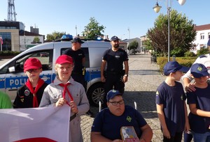 Na zdjęciu zuchy i harcerze trzymający flagę Polski w tle dwóch policjantów w umundurowaniu służbowym i radiowóz oraz budynki otaczające lubaczowski rynek.