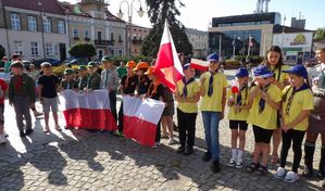 Na zdjęciu zuchy i harcerze trzymający flagę Polski w tle budynki otaczjące rynek.