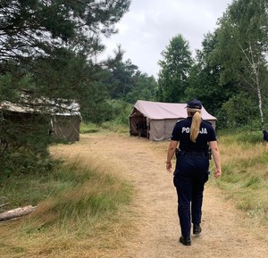 Na zdjęciu policjantka w umundurowaniu służbowym w tle las i namioty.