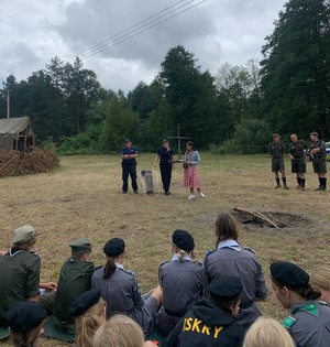 Na pierwszym planie słuchacze w środku stoją policjanci i harcerze w tle las i namioty.