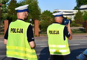 Na zdjęciu dwóch policjantów umundurowanych w kamizelkach odblaskowych, którzy dokonują pomiaru prędkości.