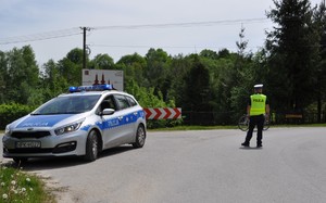 Na zdjęciu droga na której stoi policjant w umundurowaniu służbowym i kamizelce oraz radiowóz. W tle zabudowania i drzewa.