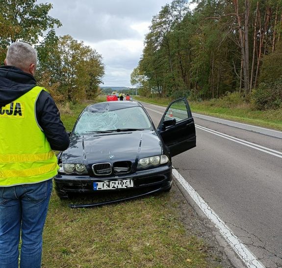 Na drodze stoi rozbity pojazd osobowy marki BMW, policjant w kamizelce odblaskowej roi zdjęcie, w tle służby ratunkowe.