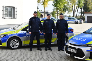 Trzech umundurowanych policjantów za nimi radiowóz i budynek komendy.