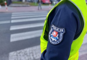 Na tle przejścia dla pieszych i budynków widać tor policjanta i rękę w umundurowaniu służbowym i kamizelce odblaskowej, na rękawie emblemat z logiem i napisem Komenda Powiatowa Policji w Lubaczowie.