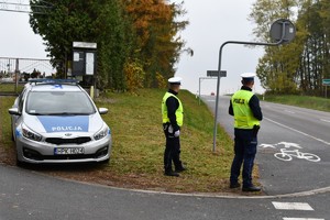 Na zdjęciu dwóch policjantów w umundurowaniu służbowym. Stoją przy drodze i radiowozie. W tle cmentarz.