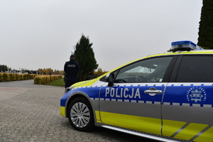 Oznakowany radiowóz policyjny, przed nim policjant w umundurowaniu służbowym w tle cmentarz i niebo.